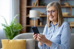 ein mittleren Alters Frau im ein Blau Hemd Sitzung auf ein Couch, absorbiert im ihr Smartphone im ein gut beleuchtet, gemütlich Leben Zimmer Einstellung. foto