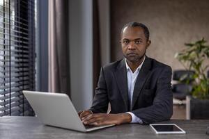 konzentriert afrikanisch amerikanisch Geschäftsmann im ein dunkel passen funktioniert von seine Laptop im ein gut beleuchtet modern Büro, ausströmend Vertrauen und Sachverstand. foto