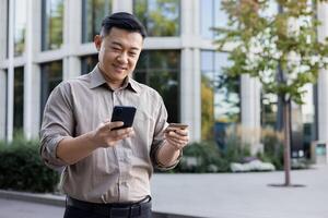 ein jung lächelnd asiatisch Mann ist Stehen auf ein Stadt Straße, halten ein Anerkennung Karte und mit ein Handy, Mobiltelefon Telefon. foto