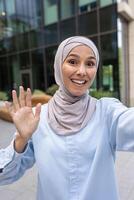 ein heiter Frau im ein Hijab ist winken während nehmen ein Vertikale Selfie, scheinbar auf ein Forderung, inmitten ein städtisch Hintergrund. foto