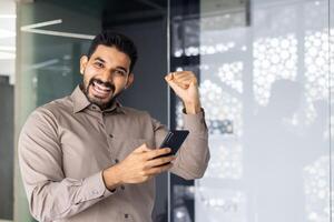 ein heiter Mann im ein beiläufig Hemd feiert Erfolg mit ein angehoben Faust im ein Büro Einstellung, halten seine Telefon. foto