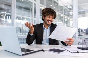 jung erfolgreich Financier Investor Innerhalb Büro beim Arbeitsplatz mit Unterlagen und Laptop, spanisch Mann im Geschäft passen suchen beim Berichte und Grafiken, Geschäftsmann zufrieden mit Leistung Ergebnisse. foto