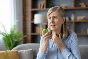 krank Senior Dame berühren Hals und Überschrift Aerosol zu Mund während Gefühl schwer zu Atem im Wohnung Innere. Asthmatiker weiblich mit verschreiben Behandlung mit Analgetikum während Allergie Attacke. foto