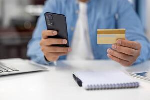 Nahansicht von ein männlich Hände mit ein Smartphone und Anerkennung Karte zum ein online Transaktion auf ein Weiß Schreibtisch mit ein Notizblock. foto