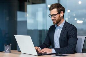 erfolgreich reifen Geschäftsmann beim Arbeit mit Laptop Innerhalb Büro, Mann im Geschäft passen Sitzung beim Schreibtisch Tippen auf Computer, zufrieden mit Leistung Ergebnis und arbeiten. foto