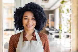 Porträt von Brünette vielfältig lächelnd Dame tragen Schürze Über Bluse während lächelnd beim Kamera auf verschwommen Hintergrund. optimistisch Kellnerin Begrüßung Kunden im leeren Restaurant während Tageszeit. foto