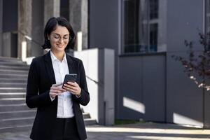 zuversichtlich weiblich Exekutive im ein passen beschäftigt, verlobt mit ihr Smartphone draußen ein Büro Gebäude, verkörpern Professionalität und Technologie. foto