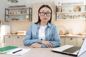 Porträt von ein jung asiatisch Geschäftsfrau Arbeiten beim Zuhause und Dirigieren online Interviews von ein Laptop. ein ernst, zielgerichtet Frau ist Sitzung beim das Tisch, suchen beim das Kamera. foto