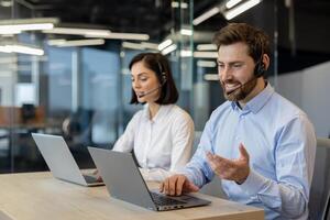 kaukasisch weiblich und männlich im korporativ Kleid Code Kleider und Kopfhörer mit Mikro Sitzung durch tragbar Computers. zwei Kunde Pflege Arbeitskräfte führen Konversation mit Kunden im modern Büro. foto