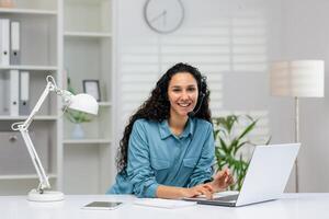 ein heiter Frau im ein Blau Hemd und tragen ein Headset Arbeiten auf ein Laptop im ein hell, organisiert Zuhause Büro Einstellung. sie erscheint beschäftigt, verlobt und freundlich. foto