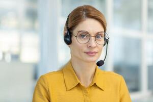 Porträt von ein jung weiblich Lehrer tragen Brille und tragen ein Headset. Sitzung im das Büro und suchen beim das Kamera. führt online Entfernung Lernen. Nahansicht Foto. foto