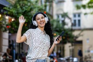 schön jung Latein amerikanisch Frau Spaziergänge im das Abend Stadt auf ein Reise, Frau mit Kopfhörer hört zu zu Musik- und Tänze und singt, Verwendet ein online Radio Anwendung auf ihr Telefon foto