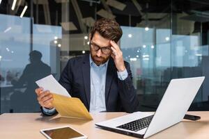 traurig und verärgert reifen Geschäftsmann empfangen ein Briefumschlag durch Mail mit ein Schlecht Nachrichten Benachrichtigung Brief, Boss im Geschäft passen Arbeiten Innerhalb Büro mit Laptop tragen Brille und Bart. foto