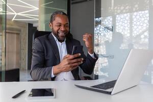 reifen erfolgreich afrikanisch amerikanisch Geschäftsmann empfangen online Benachrichtigung von gewinnen Botschaft beim Arbeitsplatz Innerhalb Büro, Mann halten Telefon, mit App auf Smartphone, glücklich Gewinner mit Leistung. foto