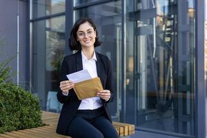 ein Fachmann Geschäftsfrau halten ein Briefumschlag und Unterlagen, lächelnd selbstbewusst während Sitzung draußen ein zeitgenössisch Büro Gebäude. foto