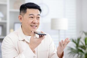 Nahansicht Foto von ein jung lächelnd asiatisch Mann Sitzung beim Zuhause reden auf das Telefon durch das Freisprecheinrichtung, Aufzeichnung das Gespräch, fragen im das Anwendung.