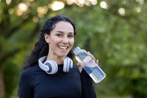 froh sportlich Frau ruhen und Trinken Wasser im Park mit Kopfhörer um ihr Nacken, lächelnd beim das Kamera. foto