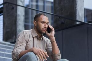 ein ernst und schockiert spanisch afrikanisch amerikanisch Mann. er sitzt auf das Schritte, Gespräche auf das Telefon traurig foto