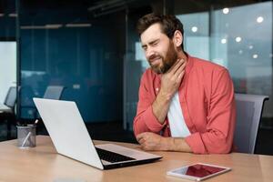 ein Mann mit ein wund Kehle funktioniert Innerhalb das Büro, ein Geschäftsmann beim Arbeit hat ein wund Kehle Grippe und kalt, Verwendet ein Laptop beim arbeiten. foto