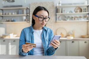 verärgert asiatisch Frau versuchen zu machen ein Kauf im ein online online speichern, Frau beim Zuhause Sitzung auf ein Sofa im das Küche mit ein Anwendung auf ein Smartphone und halten ein Bank Anerkennung Karte foto