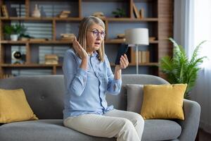 reifen Dame zeigen Verwechslung und Enttäuschung während mit ihr Handy, Mobiltelefon Telefon, Sitzung auf das Couch im ein gemütlich Leben Zimmer. foto