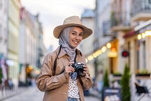 Porträt von jung schön Muslim Frau im Hijab und Hut, Tourist Gehen im Abend Stadt mit Kamera, Frau Reisen zu anders Länder. foto