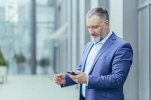 ein ernst und konzentriert Senior männlich Geschäftsmann ist Stehen in der Nähe von ein Büro Center, halten ein Handy, Mobiltelefon Telefon und ein Anerkennung Karte im seine Hände, eintreten das Karte Nummer in das Konto auf das Telefon. foto