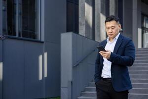 konzentriert asiatisch Geschäftsmann im ein passen Gehen draußen Büro Gebäude, vertieft im seine Smartphone Gerät. foto