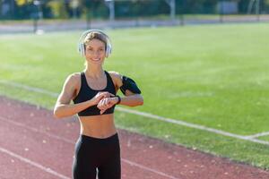 aktiv jung Frau tragen Sportbekleidung prüft ihr Smartwatch während genießen Musik- auf ihr Kopfhörer beim ein Spur Feld. foto