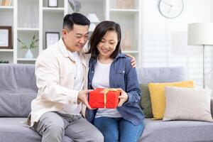 glücklich jung asiatisch Paar, Mann und Frau Sitzung auf Sofa beim heim. ein Mann gibt ein Frau ein rot Box wie ein Überraschung Geschenk. Sie Lächeln beim jeder andere. foto