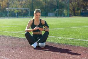 aktiv jung Frau genießt ein brechen auf ein Sport Schiene, wählen Musik- auf ihr Telefon mit Kopfhörer auf ein hell Tag. foto