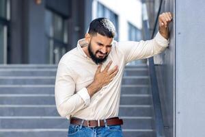 Panik und Herz Attacke auf das Straße. ein jung indisch Mann ist Stehen in der Nähe von ein Gebäude, gelehnt gegen ein Mauer und halten seine Truhe mit seine Hand, Gefühl stark Schmerzen und brauchen medizinisch Hilfe foto