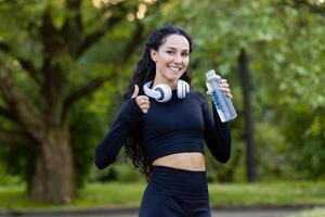 ein heiter spanisch Frau genießt ein Fitness brechen beim das Park, halten ein Wasser Flasche und tragen Kopfhörer, geben ein Daumen oben mit Bäume im das Hintergrund. foto