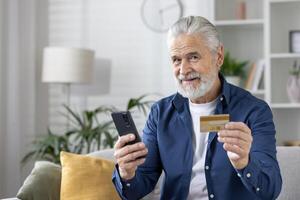 lächelnd Alten Mann halten ein Anerkennung Karte und mit ein Handy, Mobiltelefon Telefon im ein gemütlich Leben Zimmer Einstellung, vorbildlich online Einkaufen oder Banken. foto