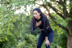 Porträt von ein schön weiblich Athlet im das Park, spanisch Frau ruht und atmet während Joggen und aktiv physisch Übung, weiblich Athlet im Sportbekleidung hat Truhe Schmerz. foto