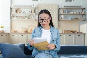 ein jung schön asiatisch Schüler Mädchen empfangen ein Universität Eintritt Brief im das Post. aufgeregt, sie hält ein Briefumschlag im ihr Hände. Sitzung beim Zuhause auf das Couch, Lektüre. foto