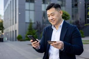 zuversichtlich asiatisch männlich Exekutive Handhabung finanziell Transaktionen mit ein Anerkennung Karte und Handy, Mobiltelefon Telefon draußen modern Büro Gebäude. foto