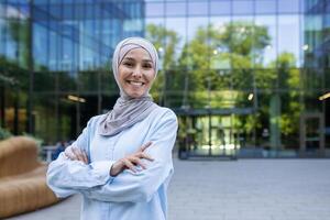 ein Fachmann Frau im ein Hijab steht selbstbewusst mit gekreuzt Waffen draußen ein zeitgenössisch Büro Gebäude, symbolisieren Ermächtigung und Vielfalt im das Arbeitsplatz. foto