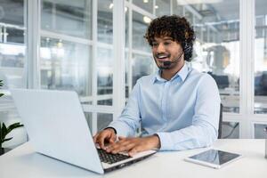 heiter männlich Kunde Bedienung Vertreter mit Headset Arbeiten auf Laptop im ein hell, zeitgenössisch Büro Einstellung. Fachmann Kommunikation und Unterstützung. foto