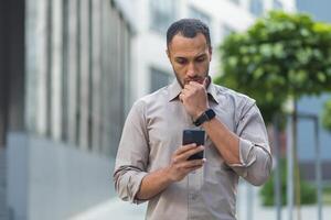 Denken und traurig Muslim Geschäftsmann lesen Botschaft von Smartphone, Mann draußen Büro Gebäude mit Telefon deprimiert lesen Schlecht Nachricht. foto