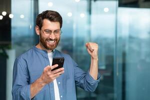 reifen Anfang Unternehmer mit Telefon in der Nähe von Fenster Innerhalb modern Büro Gebäude, Geschäftsmann mit Bart und Brille feiern Sieg und Triumph, Mann im Hemd empfangen gut Nachrichten online. foto