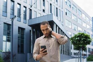 nachdenklich und aufgeregt afrikanisch amerikanisch Arbeiter draußen Büro Gebäude lesen Schlecht Nachricht, Mann halten Telefon foto