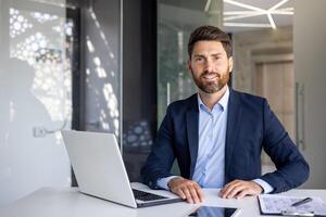 Porträt von ein jung erfolgreich und lächelnd männlich Anwalt und Politiker Sitzung im das Büro beim das Tabelle mit ein Laptop und suchen ,stolz beim das Kamera. foto