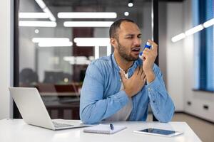 ein Mann von indisch Abstammung ist im ein Büro Einstellung, mit ein Asthma Inhalator, zeigen Zeichen von Atemwege Not. foto