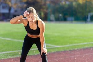 müde Sportlerin ruhen nach Joggen auf ein sonnig Tag beim das Stadion, blond im Sportbekleidung nehmen ein Verschnaufpause. foto