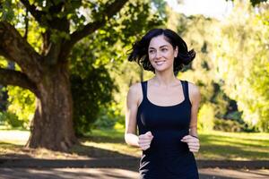 lächelnd jung schön Frau gekleidet im schwarz Sportbekleidung ist Joggen im das Park auf ein sonnig Tag. foto