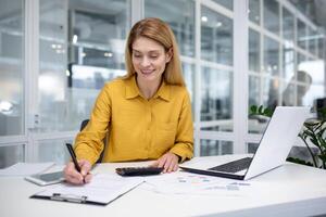 erfolgreich reifen weiblich finanziell Buchhalter beim Arbeitsplatz Innerhalb Büro, füllt Daten im Tisch, funktioniert mit Unterlagen und Papiere, Verwendet Laptop und Taschenrechner zum Papierkram. foto