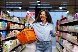 Porträt von ein glücklich Frau Käufer im ein Supermarkt, ein spanisch Frau mit ein Korb von Waren lächelt mit Vergnügen und Tänze unter das Regale mit Waren, ein zufrieden Geschäft Kunde foto