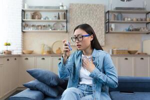 allergisch Attacke. Asthma. jung asiatisch Frau Sitzung auf Sofa beim Zuhause und mit Inhalator. hat ein Asthma Attacke, Leckereien Allergien, hat Schwierigkeit Atmung. foto