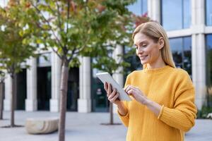 ein lächelnd reifen Frau im ein beschwingt Gelb Sweatshirt ist mit ein Digital Tablette draußen ein modern Gebäude, porträtieren Technologie verwenden und aktiv Lebensstil. foto